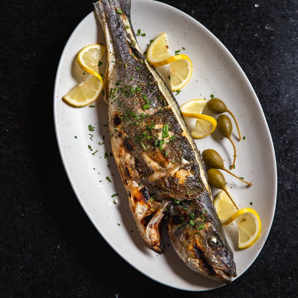 Whole grilled fish, garnished with lemon wedges, caper berries, and fresh parsley on a white plate.