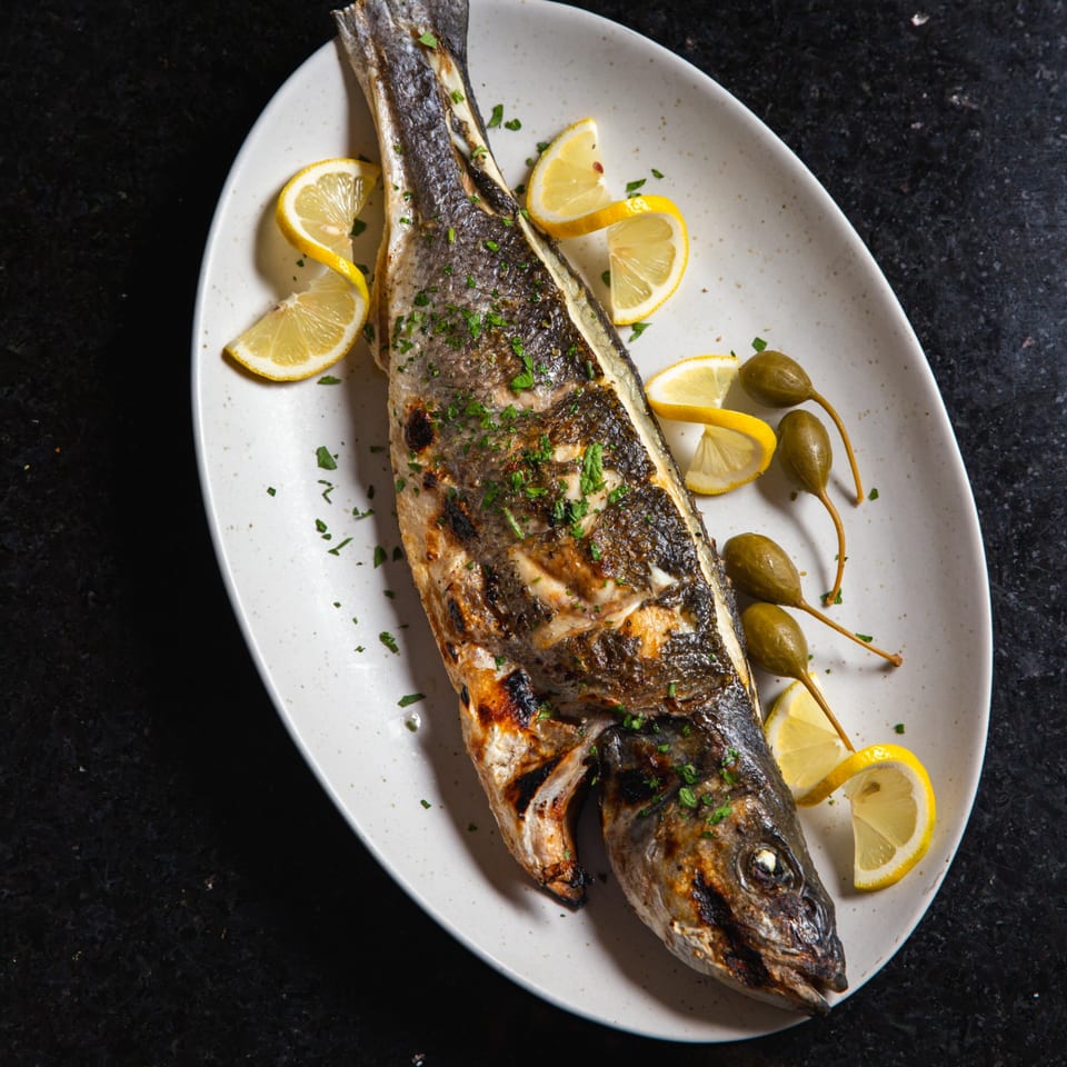 Whole grilled fish, garnished with lemon wedges, caper berries, and fresh parsley on a white plate.