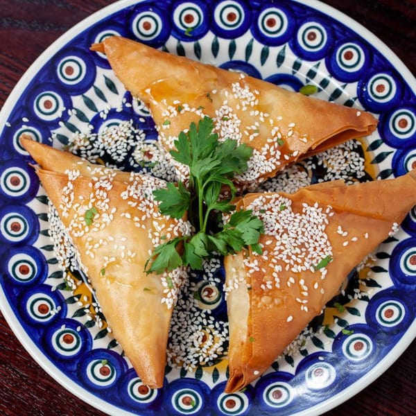 Three golden-brown Greek pastries, drizzled with honey and sesame seeds, garnished with parsley on a blue and white plate.