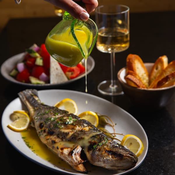 A hand pours dressing over a whole grilled fish with lemon, alongside salad, bread, and wine.