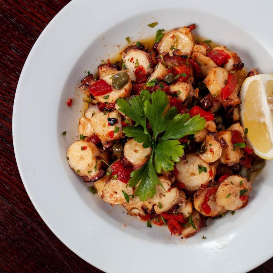 Grilled octopus with red peppers, capers, parsley, and a lemon wedge in a white bowl.