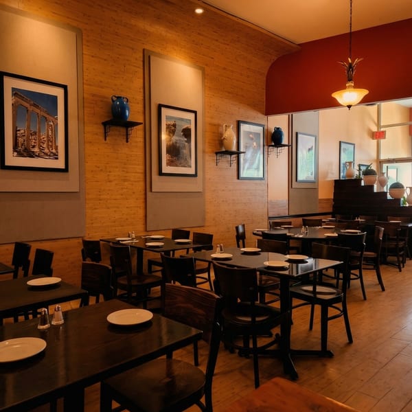 An empty restaurant dining room with dark tables, chairs, and framed artwork on light wood paneling.