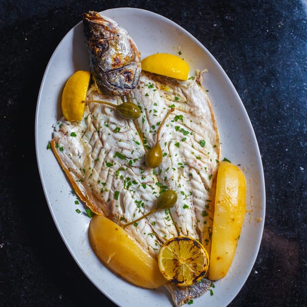 Grilled whole fish with lemon wedges, caper berries, and roasted potatoes on a white plate.