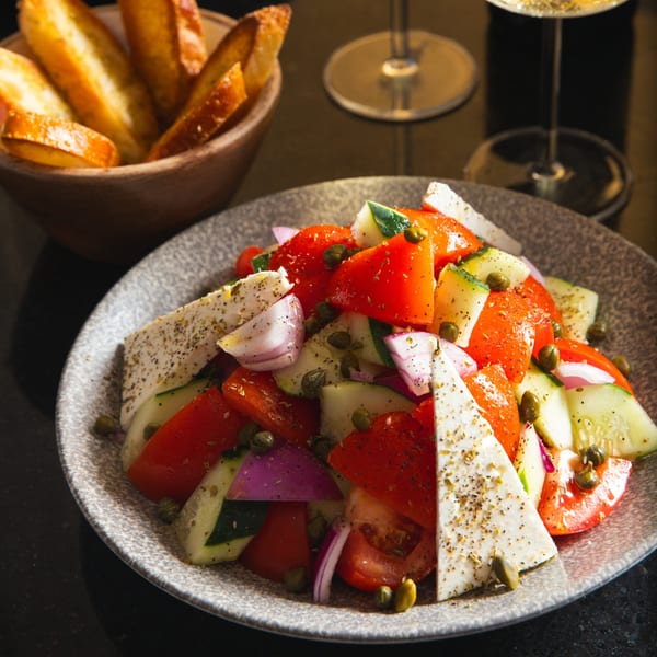 A vibrant Greek salad with tomatoes, cucumbers, red onions, feta, and capers, served with toasted bread.