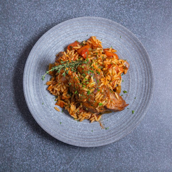 Braised lamb shank with orzo pasta, tomatoes, and herbs on a grey plate.