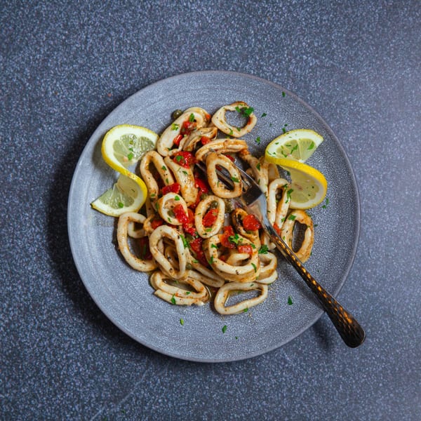 Calamari rings with red peppers, parsley, and lemon wedges on a gray plate with a fork.