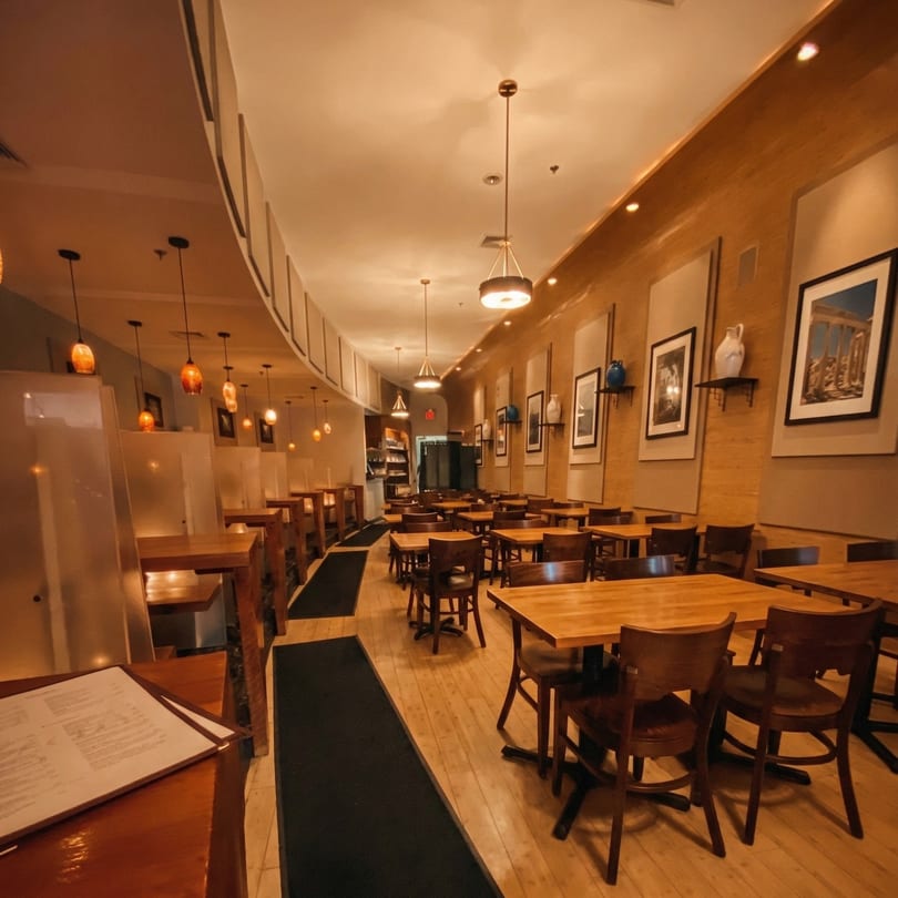 Empty restaurant dining room with wooden tables, chairs, and warm lighting, featuring framed art on the walls.