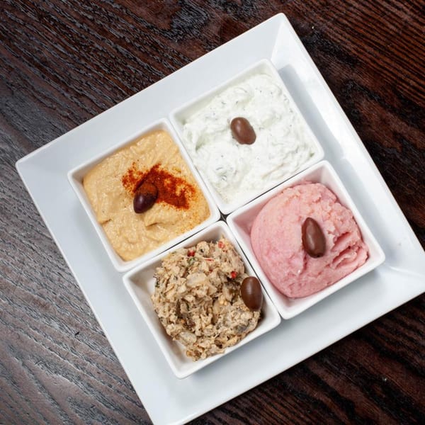 A white square platter holds four small dishes of Greek dips, each garnished with an olive.