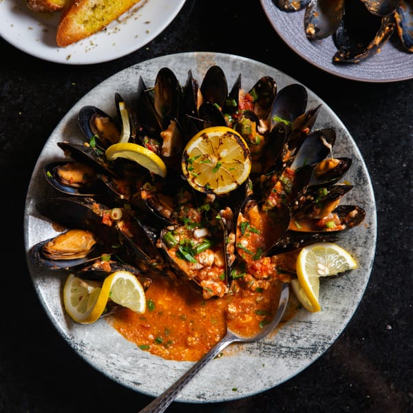 Steamed mussels in a rich red sauce, garnished with fresh herbs and lemon slices, served with garlic bread.