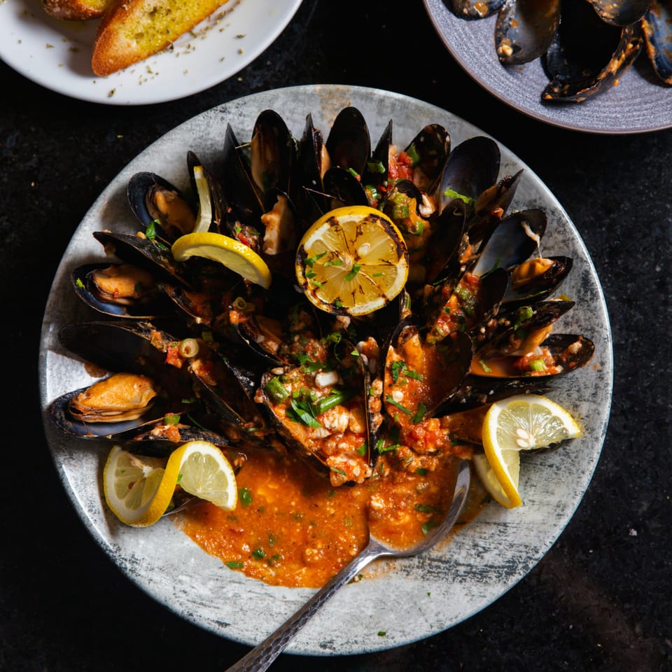 Steamed mussels in a rich red sauce, garnished with fresh herbs and lemon slices, served with garlic bread.