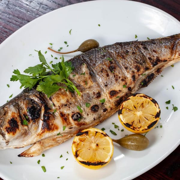 Grilled whole fish with charred lemon halves, caper berries, and fresh parsley on a white plate.