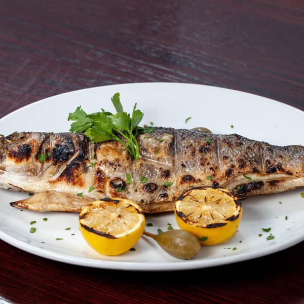 Whole grilled fish with charred skin, garnished with parsley, grilled lemon halves, and a caper berry.