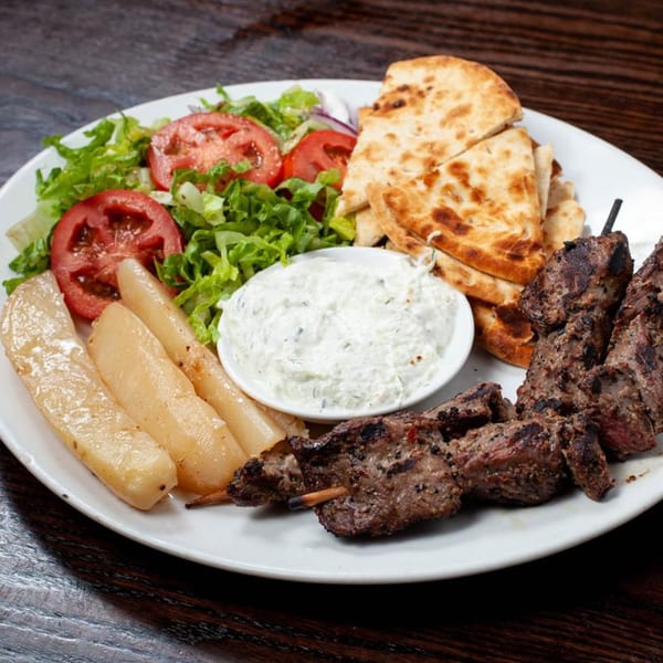 Grilled meat skewers, pita bread, salad, potatoes, and tzatziki sauce on a white plate.