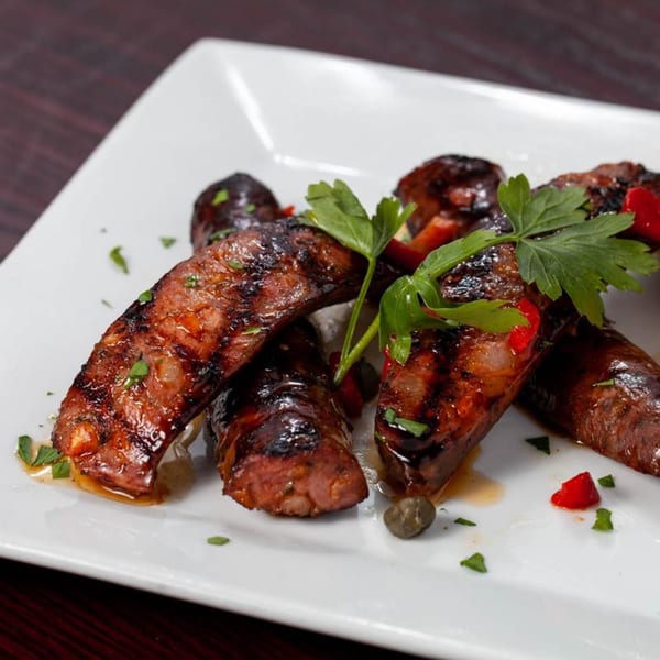 Grilled Greek sausages garnished with fresh parsley, red peppers, and capers on a white plate.