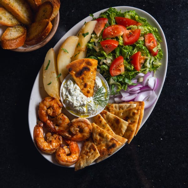 A platter with grilled shrimp, pita bread, tzatziki, roasted potatoes, and a fresh salad.