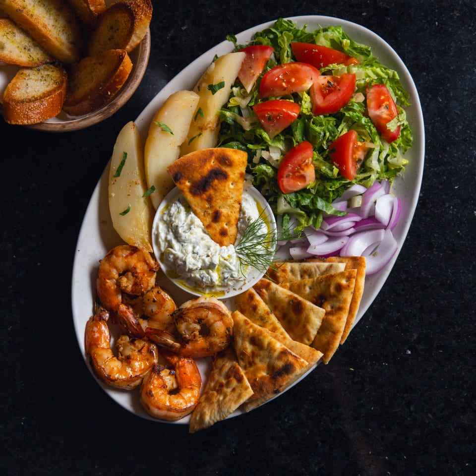 A platter with grilled shrimp, pita bread, tzatziki, roasted potatoes, and a fresh salad.