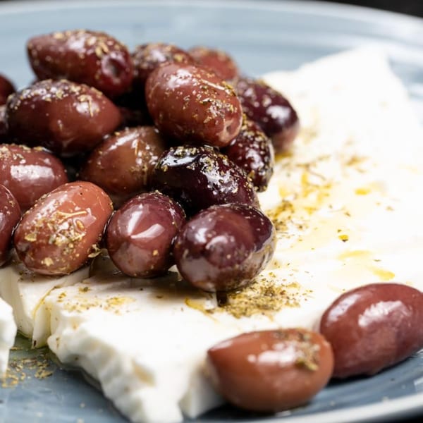 Close-up of Kalamata olives and feta cheese drizzled with olive oil and herbs on a blue plate.